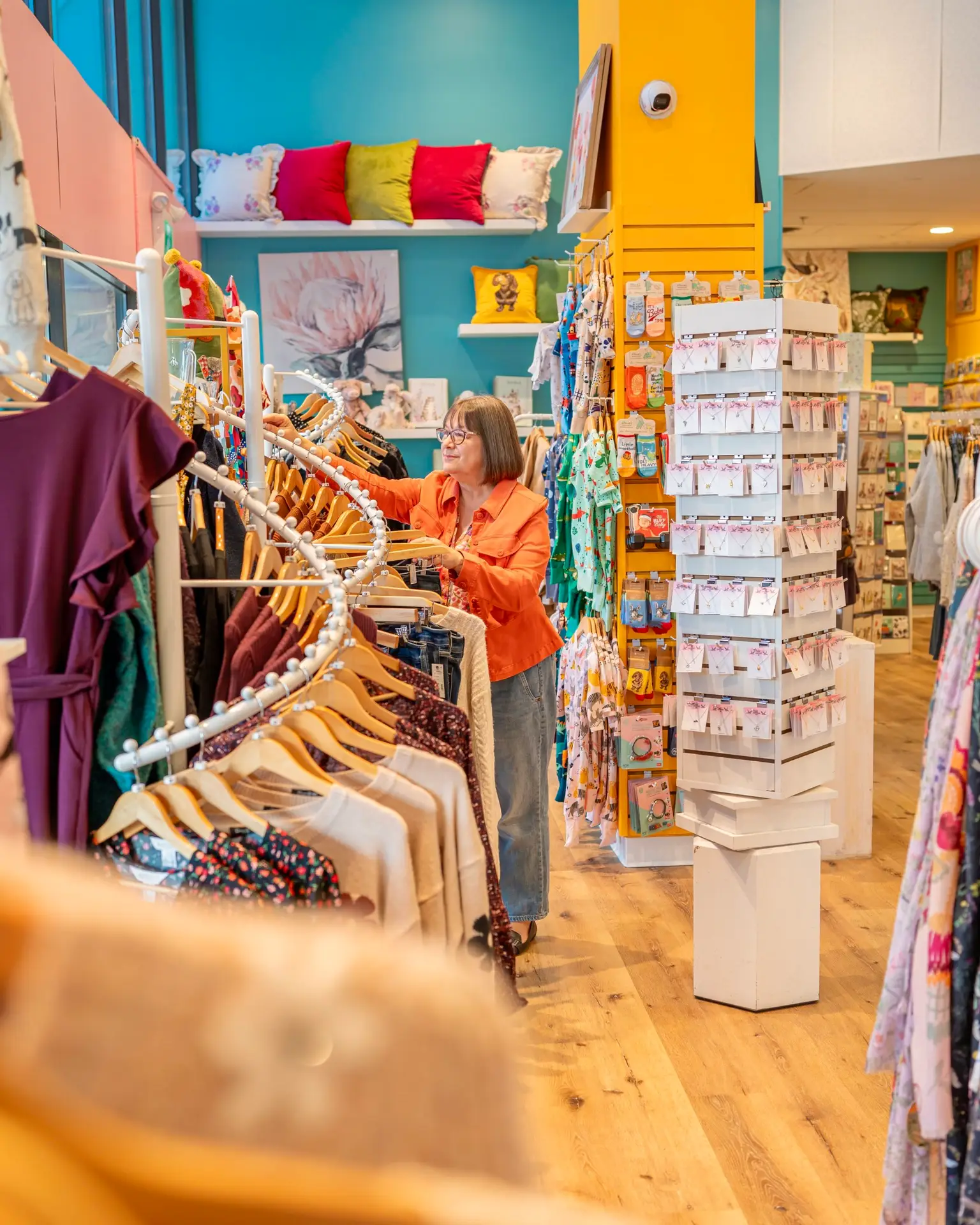 A woman browses a rack of colorful clothing inside Cameron Rose Gifts in Sidney, BC, surrounded by bright teal and yellow walls, patterned pillows, gift displays, and neatly arranged accessories.